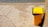 A yellow garbage bin with a black liner is positioned next to a wall on a paved surface. The pavement is cracked and weathered, and the bin casts a shadow on the ground. In the background, there is a section of rough, pebbled texture, possibly indicating a beach or riverside.