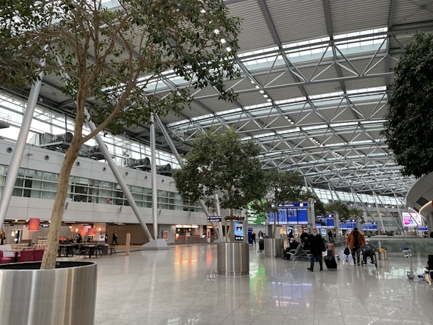 A spacious airport terminal with a high ceiling featuring metal beams and large windows allowing natural light. There are several trees in planters along the walkway, with people walking and some sitting near dining areas. Flight information boards are visible in the background.
