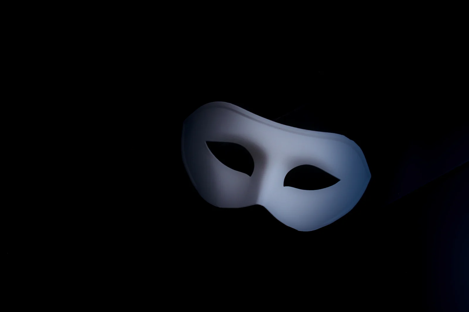 A plain white masquerade-style eye mask resting in near-total darkness, lit from one side