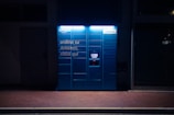 A blue Amazon parcel locker is illuminated against a dark background, featuring compartments and a screen for package retrieval. It displays phrases in Italian, suggesting its use for ordering and pickup.
