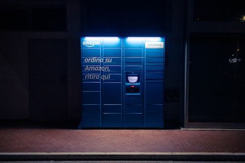 A blue Amazon parcel locker is illuminated against a dark background, featuring compartments and a screen for package retrieval. It displays phrases in Italian, suggesting its use for ordering and pickup.