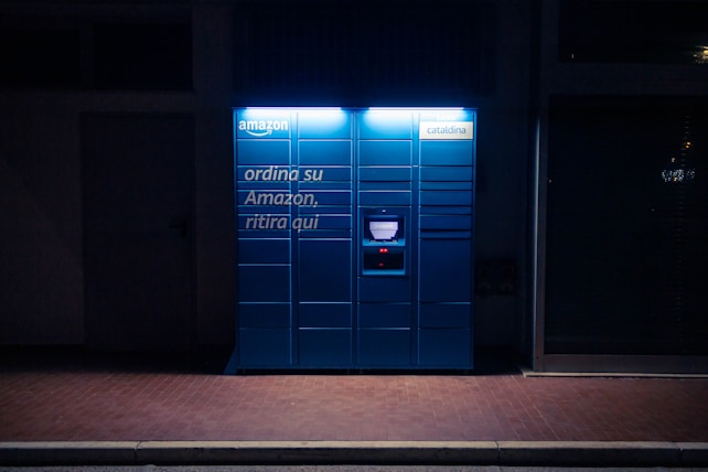 A blue Amazon parcel locker is illuminated against a dark background, featuring compartments and a screen for package retrieval. It displays phrases in Italian, suggesting its use for ordering and pickup.