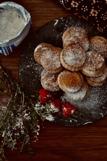 A plate filled with round, flaky pastries dusted with powdered sugar is placed on a wooden table. To the side, there are fresh strawberries, also lightly dusted with sugar. A small bag of flour and some fresh herbs are visible nearby.