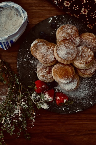 A plate filled with round, flaky pastries dusted with powdered sugar is placed on a wooden table. To the side, there are fresh strawberries, also lightly dusted with sugar. A small bag of flour and some fresh herbs are visible nearby.