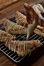 A hand is piping white icing in a zigzag pattern onto freshly baked puff pastries. The pastries are placed on a metal cooling rack over a wooden table. The pastries are golden brown with a flaky texture, and the icing adds a decorative finishing touch.