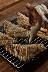 A hand is piping white icing in a zigzag pattern onto freshly baked puff pastries. The pastries are placed on a metal cooling rack over a wooden table. The pastries are golden brown with a flaky texture, and the icing adds a decorative finishing touch.
