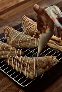 A hand is piping white icing in a zigzag pattern onto freshly baked puff pastries. The pastries are placed on a metal cooling rack over a wooden table. The pastries are golden brown with a flaky texture, and the icing adds a decorative finishing touch.