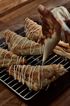 A hand is piping white icing in a zigzag pattern onto freshly baked puff pastries. The pastries are placed on a metal cooling rack over a wooden table. The pastries are golden brown with a flaky texture, and the icing adds a decorative finishing touch.