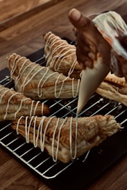 A hand is piping white icing in a zigzag pattern onto freshly baked puff pastries. The pastries are placed on a metal cooling rack over a wooden table. The pastries are golden brown with a flaky texture, and the icing adds a decorative finishing touch.