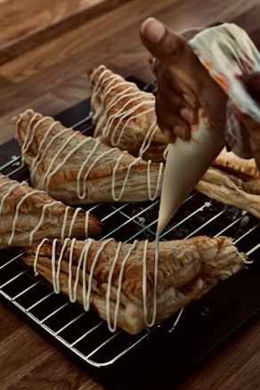 A hand is piping white icing in a zigzag pattern onto freshly baked puff pastries. The pastries are placed on a metal cooling rack over a wooden table. The pastries are golden brown with a flaky texture, and the icing adds a decorative finishing touch.