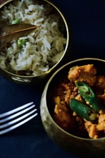 Two brass bowls are filled with Indian cuisine; one contains chicken curry garnished with green chilies, while the other holds rice mixed with green peas and topped with a bay leaf. A metal fork lies beside the bowls on a dark surface.