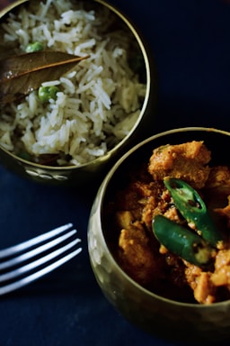 Two brass bowls are filled with Indian cuisine; one contains chicken curry garnished with green chilies, while the other holds rice mixed with green peas and topped with a bay leaf. A metal fork lies beside the bowls on a dark surface.