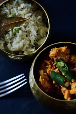 Two brass bowls are filled with Indian cuisine; one contains chicken curry garnished with green chilies, while the other holds rice mixed with green peas and topped with a bay leaf. A metal fork lies beside the bowls on a dark surface.
