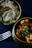 Two brass bowls are filled with Indian cuisine; one contains chicken curry garnished with green chilies, while the other holds rice mixed with green peas and topped with a bay leaf. A metal fork lies beside the bowls on a dark surface.