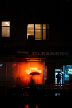 A night scene in an urban environment with a brightly lit neon sign reading 'Young Cleaners' glowing in orange, reflecting on the wet ground. A person holding an umbrella stands near the entrance of a building, partially obscured by the sign. Nearby windows are illuminated with warm light, adding to the ambiance.