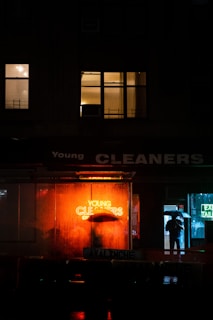 A night scene in an urban environment with a brightly lit neon sign reading 'Young Cleaners' glowing in orange, reflecting on the wet ground. A person holding an umbrella stands near the entrance of a building, partially obscured by the sign. Nearby windows are illuminated with warm light, adding to the ambiance.