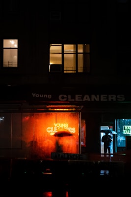 A night scene in an urban environment with a brightly lit neon sign reading 'Young Cleaners' glowing in orange, reflecting on the wet ground. A person holding an umbrella stands near the entrance of a building, partially obscured by the sign. Nearby windows are illuminated with warm light, adding to the ambiance.