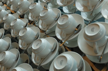 A large number of white teacups arranged neatly and uniformly, each placed upside down on saucers. Spoons are placed beside each cup in an organized manner.