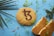 A round biscuit with a symbol resembling 't3' in brown is placed on a blue surface. The biscuit is surrounded by colorful sprinkles, a cut piece of a similar biscuit, and various small green leaves. Palm leaves are partially visible on the left.