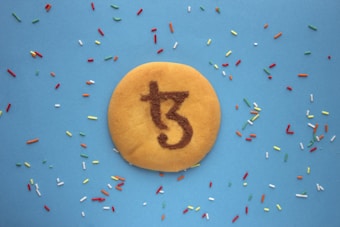 A round cookie is placed on a blue background, featuring a decorative design on its surface that resembles an abstract symbol or letter. Scattered around the cookie are colorful sprinkles in green, red, orange, white, and yellow hues.