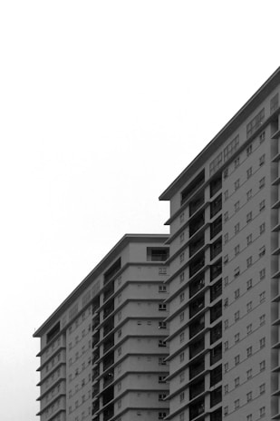 Two tall, modern apartment buildings with numerous windows, pictured in black and white, rise against a plain sky.
