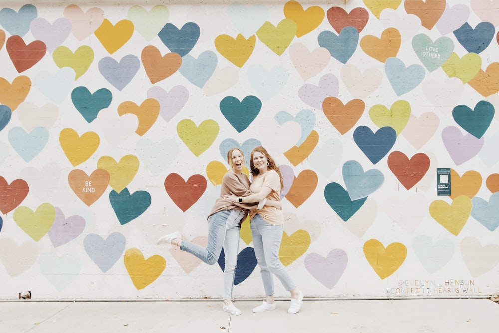 two women standing in front of a wall with hearts painted on it