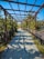 A paved path is shaded by a metal pergola structure with plants and vines growing along its sides. The pathway is surrounded by lush greenery and decorative wooden fences, creating a serene and inviting atmosphere. Sunlight casts intricate shadows on the blue-gray pavers.
