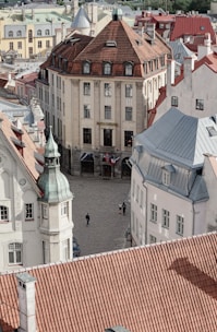 Aerial view of an urban area featuring several historical buildings with red-tiled roofs. The architecture is characterized by intricate designs and varying heights. There is a cobblestone square with a few pedestrians walking. The central building is prominent with a symmetrical façade and flags near the entrance.