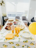 A table is set up for a gathering, adorned with a lemon-themed tablecloth and matching decor. Plates of various finger foods, stacks of disposable cups, and utensils are neatly arranged, along with lemons and a yellow balloon adding a festive touch.