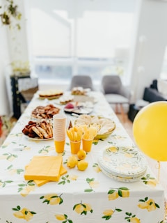 A table is set up for a gathering, adorned with a lemon-themed tablecloth and matching decor. Plates of various finger foods, stacks of disposable cups, and utensils are neatly arranged, along with lemons and a yellow balloon adding a festive touch.