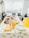 A table is set up for a gathering, adorned with a lemon-themed tablecloth and matching decor. Plates of various finger foods, stacks of disposable cups, and utensils are neatly arranged, along with lemons and a yellow balloon adding a festive touch.