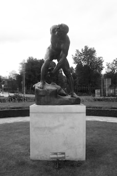 A black and white photo featuring a large bronze sculpture of a muscular, dynamic figure poised atop a stone pedestal. The setting is outdoors, surrounded by neatly trimmed grass and trees in the background. The statue has a powerful stance, emphasizing strength and movement.