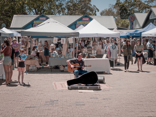 A street market scene with various stalls under white tents selling arts and crafts. A musician is playing a guitar in the center, surrounded by people walking and browsing. The backdrop consists of trees and a row of colorful traditional-style buildings.