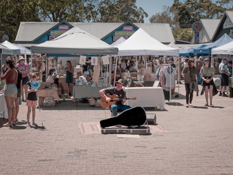 A street market scene with various stalls under white tents selling arts and crafts. A musician is playing a guitar in the center, surrounded by people walking and browsing. The backdrop consists of trees and a row of colorful traditional-style buildings.