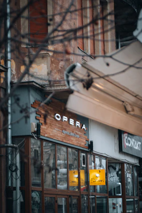 A storefront with a sign saying 'Opera', featuring large glass windows and a wooden facade. The area appears weathered and urban, with visible branches and nearby buildings.