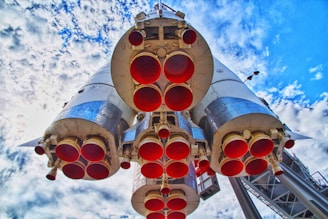 A close-up view from below of a large rocket with multiple engine nozzles facing downward. The engines are surrounded by metal casings with a reflective finish. The background is filled with a partly cloudy blue sky.