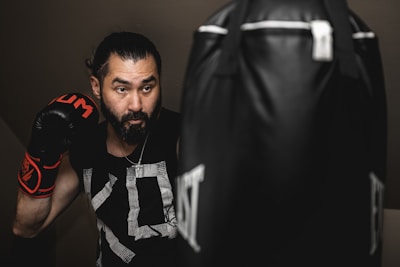A man wearing boxing gloves is practicing with a punching bag. He appears focused and determined in an indoor setting, with soft lighting creating a dramatic effect. His hair is tied back, and he's wearing a black sleeveless shirt with graphic patterns.