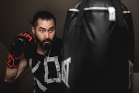 A man wearing boxing gloves is practicing with a punching bag. He appears focused and determined in an indoor setting, with soft lighting creating a dramatic effect. His hair is tied back, and he's wearing a black sleeveless shirt with graphic patterns.