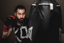 A man wearing boxing gloves is practicing with a punching bag. He appears focused and determined in an indoor setting, with soft lighting creating a dramatic effect. His hair is tied back, and he's wearing a black sleeveless shirt with graphic patterns.