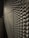 A metallic surface with a complex, repeating textured pattern that creates an optical illusion effect under low-light conditions. The surface features a series of protrusions casting shadows, adding depth to the structure.