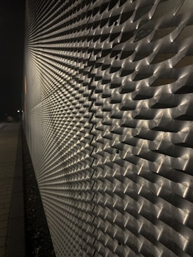 A metallic surface with a complex, repeating textured pattern that creates an optical illusion effect under low-light conditions. The surface features a series of protrusions casting shadows, adding depth to the structure.