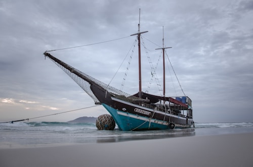A traditional wooden sailing ship is beached on the shore, with its masts towering against a cloudy sky. The ship has a distinctively painted hull, primarily in shades of black and turquoise. The sea is calm with gentle waves, and distant land is visible on the horizon.