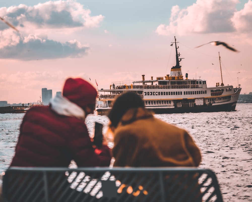 two people sitting on a bench looking at a boat