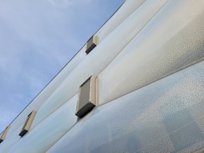The image features the facade of a modern building with a textured glass surface and several metallic ventilation units attached. The sky above is clear with a soft blue tone.
