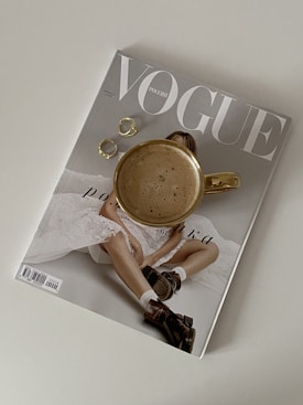 A Vogue magazine is placed on a light surface with a gold-rimmed coffee cup resting on top of it. Next to the cup are two gold rings. The magazine cover features a model in a white outfit with black footwear. The overall composition is minimalistic and elegant.