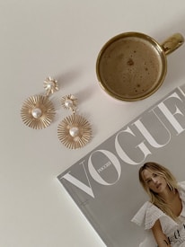 A neatly arranged flat lay featuring a cup of coffee with a golden handle, a pair of elegant gold and pearl earrings, and a Vogue magazine. The magazine cover displays a model in a white lace outfit.