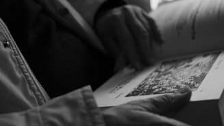 A person is holding an open book, focusing on a page with an intricate illustration. The scene is captured in black and white, highlighting the hands and the texture of the book's pages.