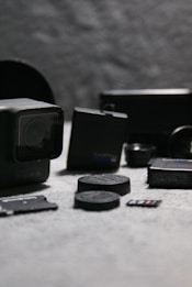 A collection of camera equipment including action cameras, battery packs, and memory cards is arranged on a grey textured surface. The foreground features a prominent action camera and memory card, with additional equipment blurred in the background.