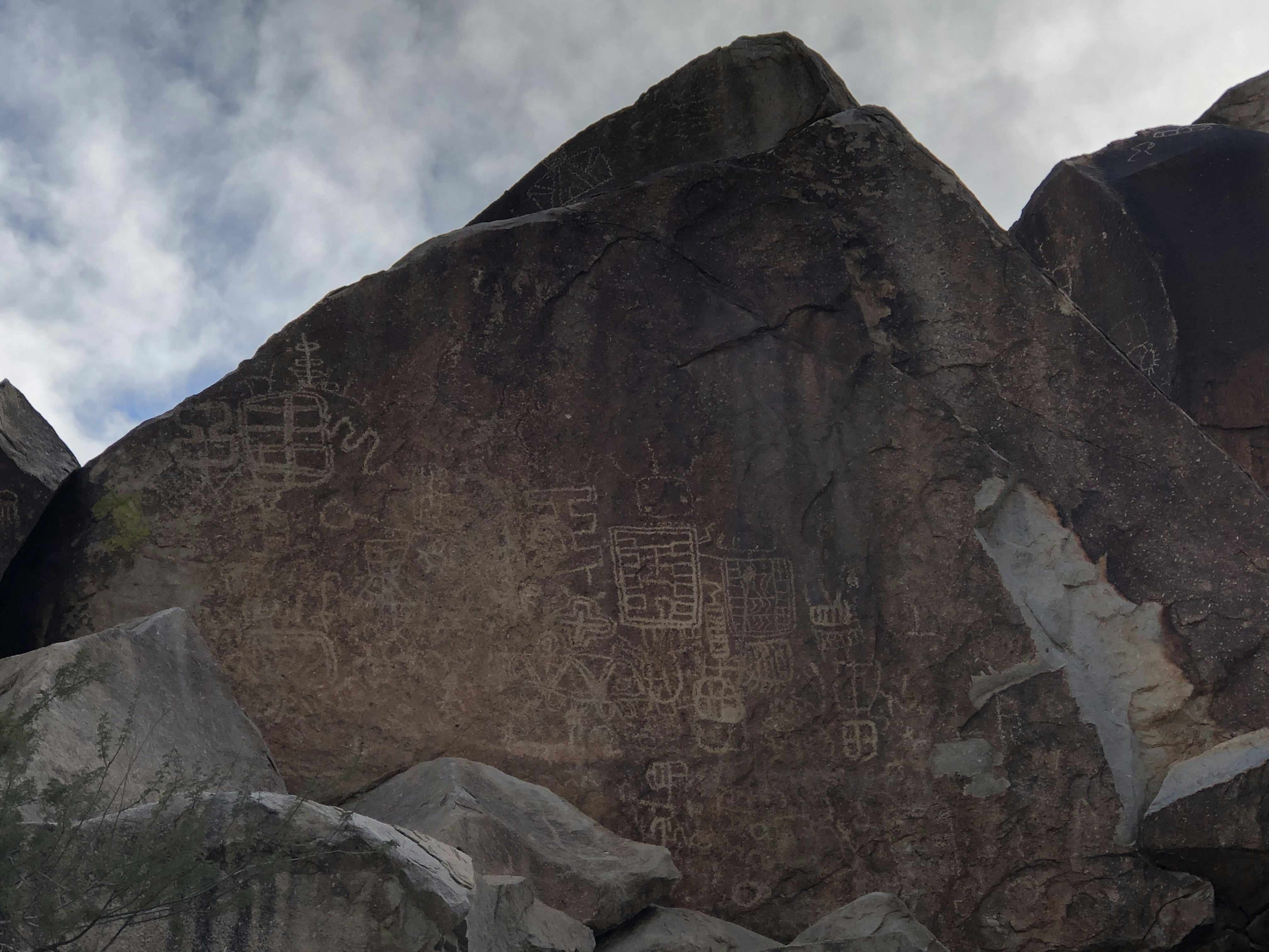 Ancient petroglyphs carved into a large rock surface, surrounded by smaller rocks, under a cloudy sky.