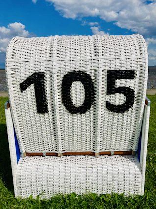 A white wicker beach chair with the number 105 displayed in black on its backrest. It is placed on a grassy area near a beach, with a body of water and partly cloudy blue sky visible in the background.